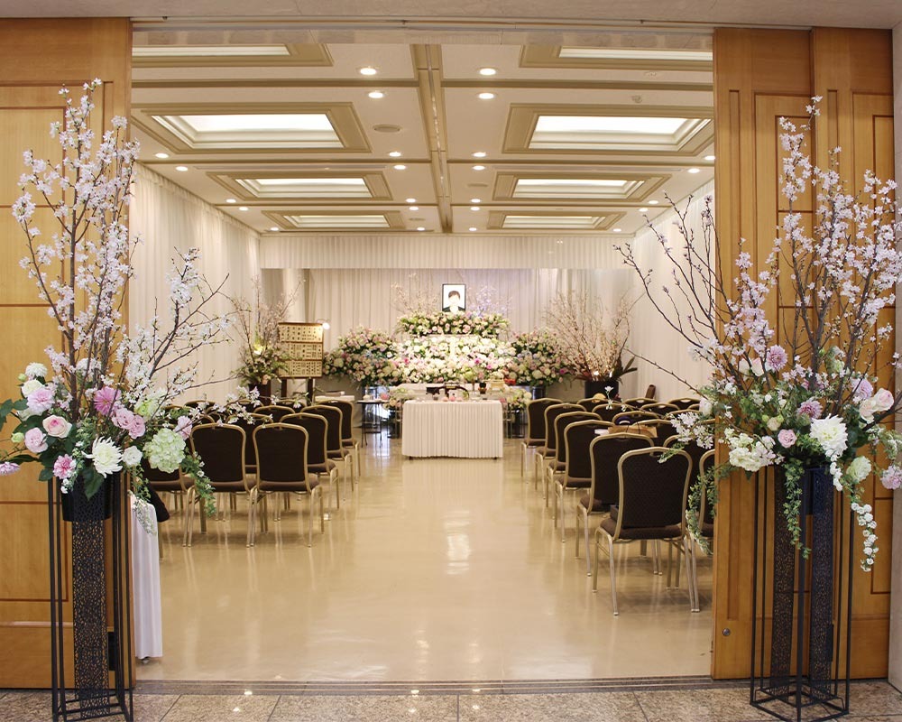 東京都品川区にある桐ヶ谷斎場での葬儀風景。たくさんのお花で囲まれた祭壇があり、式場の入り口にもピンクや白の花が備えられています。